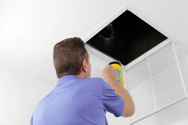 Person in a blue shirt inspects an open air vent with a flashlight, looking into the ductwork.