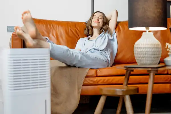 A woman in casual clothes relaxes on a brown leather couch with her feet up. A dehumidifier and a lamp on a table are in the foreground.