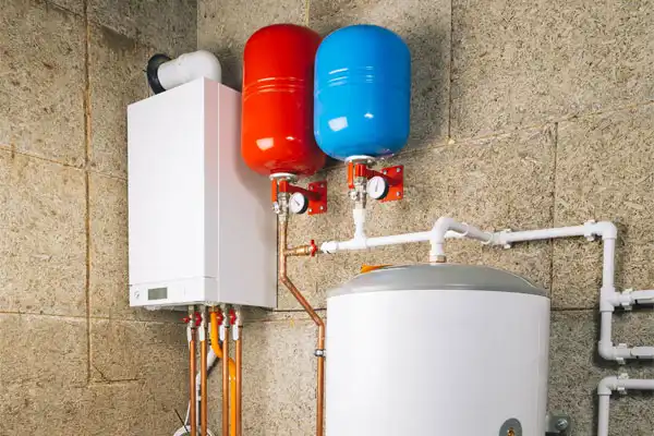 Home heating system with a wall-mounted boiler, red and blue expansion tanks, and various pipes against a textured wall.